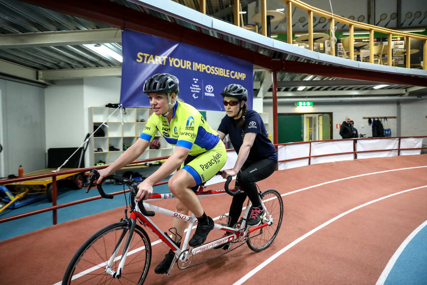 Två tjejer cyklar på en tandemcykel. Ledaren sitter längst fram på cykeln och en tjej med synnedsättning sitter bakom ledaren.