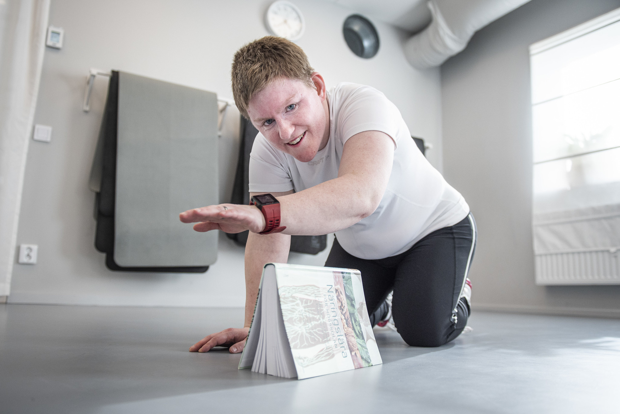 Bild på en kvinna som står på alla fyra och lyfter en arm över en bok som står placerad på golvet. Kvinnan  har en vit t-shirt och svarta träningsbyxor. I bakgrunden ser man ett fönster och träningsmattor som hänger på väggen.