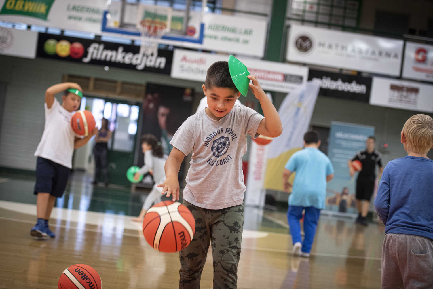 Pojke studsar en basketboll framför sig i idrottshall, andra barn studsar bollar i bakgrunden. 