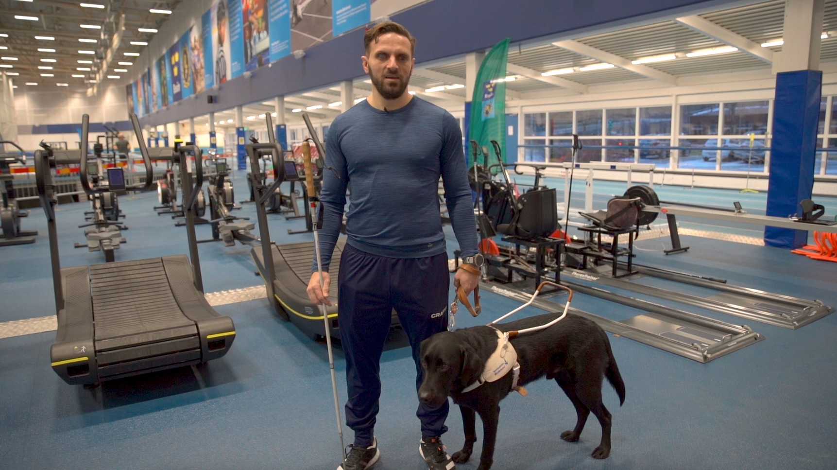 Inomhusmiljö, gym. En man med träningskläder står med kopplet till en ledarhund i ena handen och en vit käpp i andra handen. I bakgrunden är det gymutrustning.