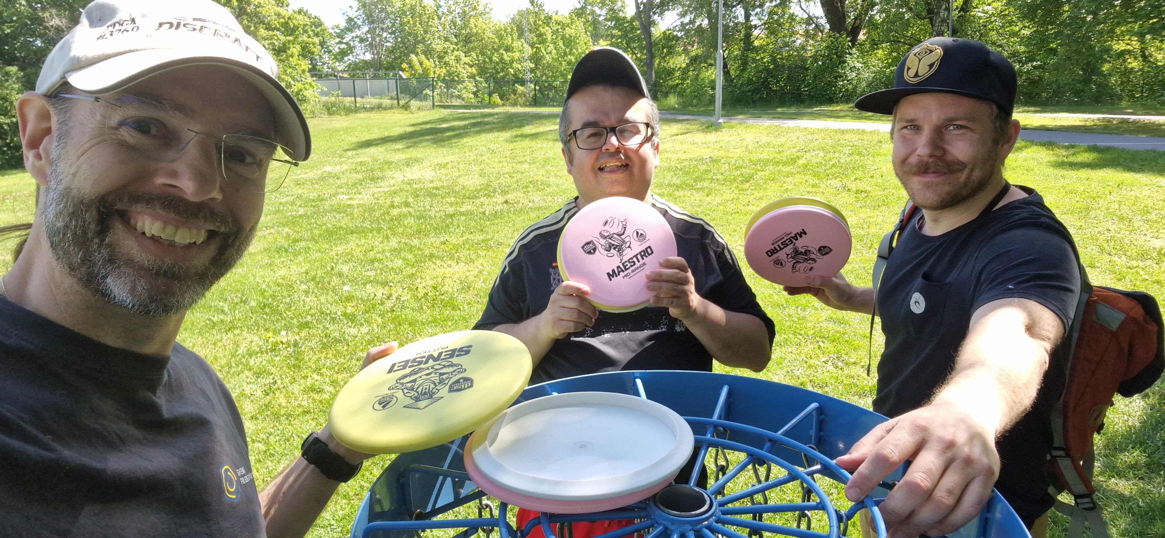 Tre personer står vid en korg och håller upp sina frisbees.