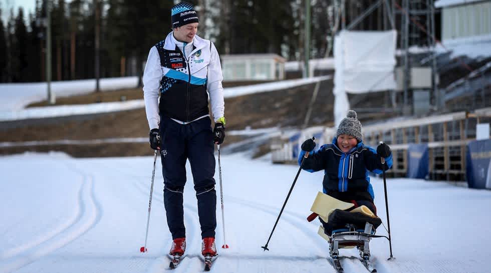 En person stående på skidor och en person i sitski åker inne på stadioområde. 