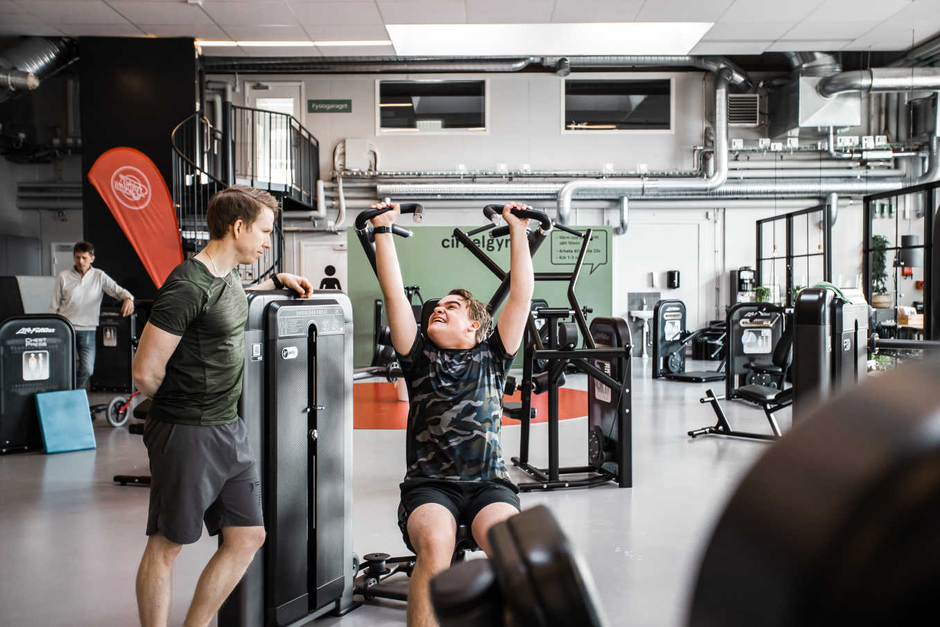 En kille tränar axelpress tillsammans med en PT på ett gym.