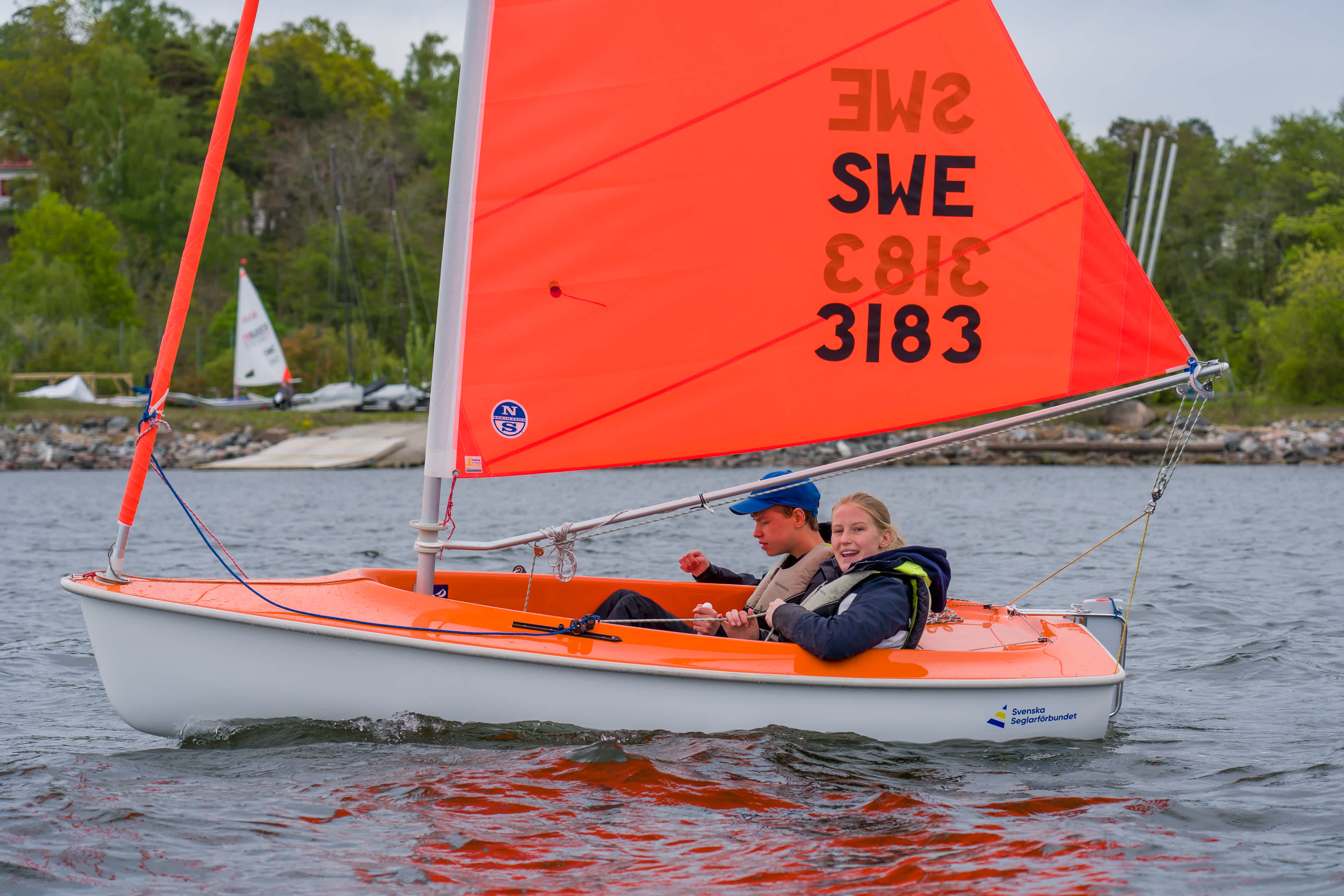 En orange segelbåt med orange segel ute på vattnet. Land med brygga och skog i bakgrunden. I båten sitter en deltagare och en ledare.