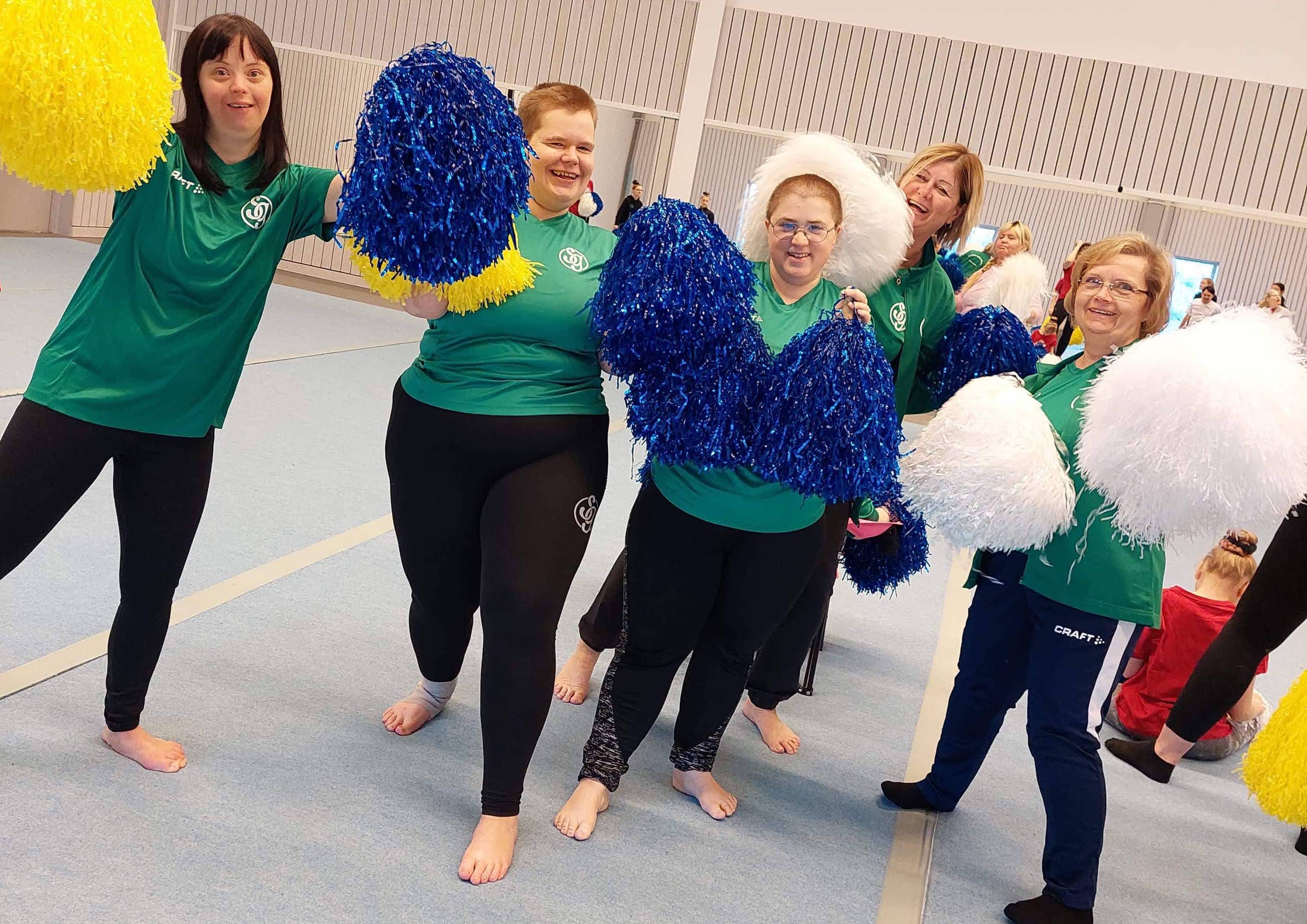 En grupp med leende gymnaster poserar med pom-poms i gymnastiksalen.