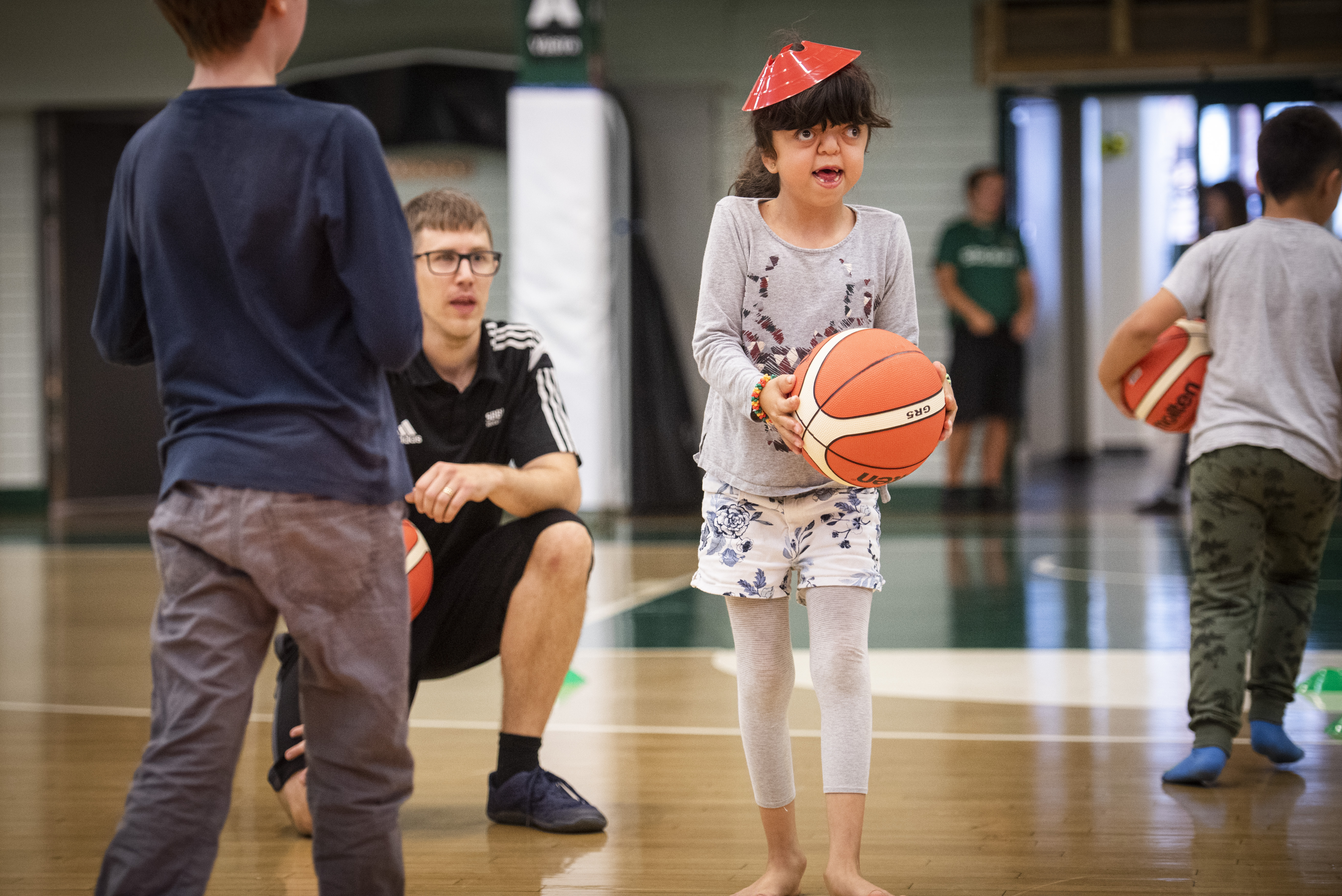 Ung flicka på med basketboll i handen. Andra barn och ledare i bakgrunden.