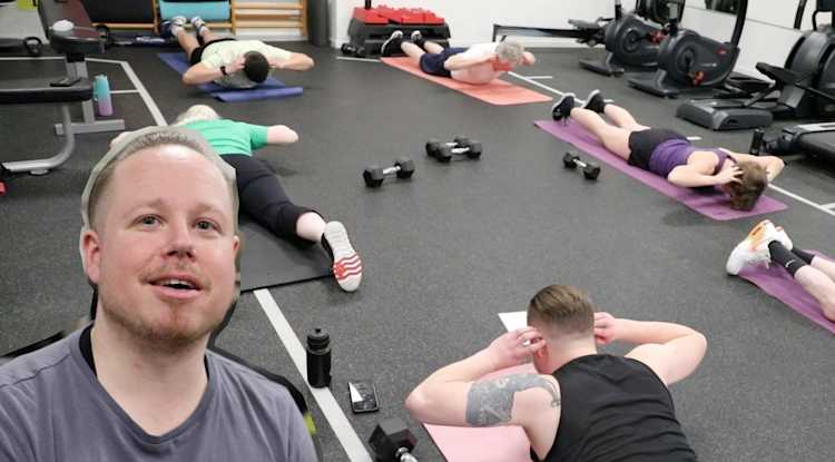 Inomhusmiljö, gym. På golvet ligger folk på träningsmattor och gör övningar. Inklippt porträttbild på Robert ifrån videon.