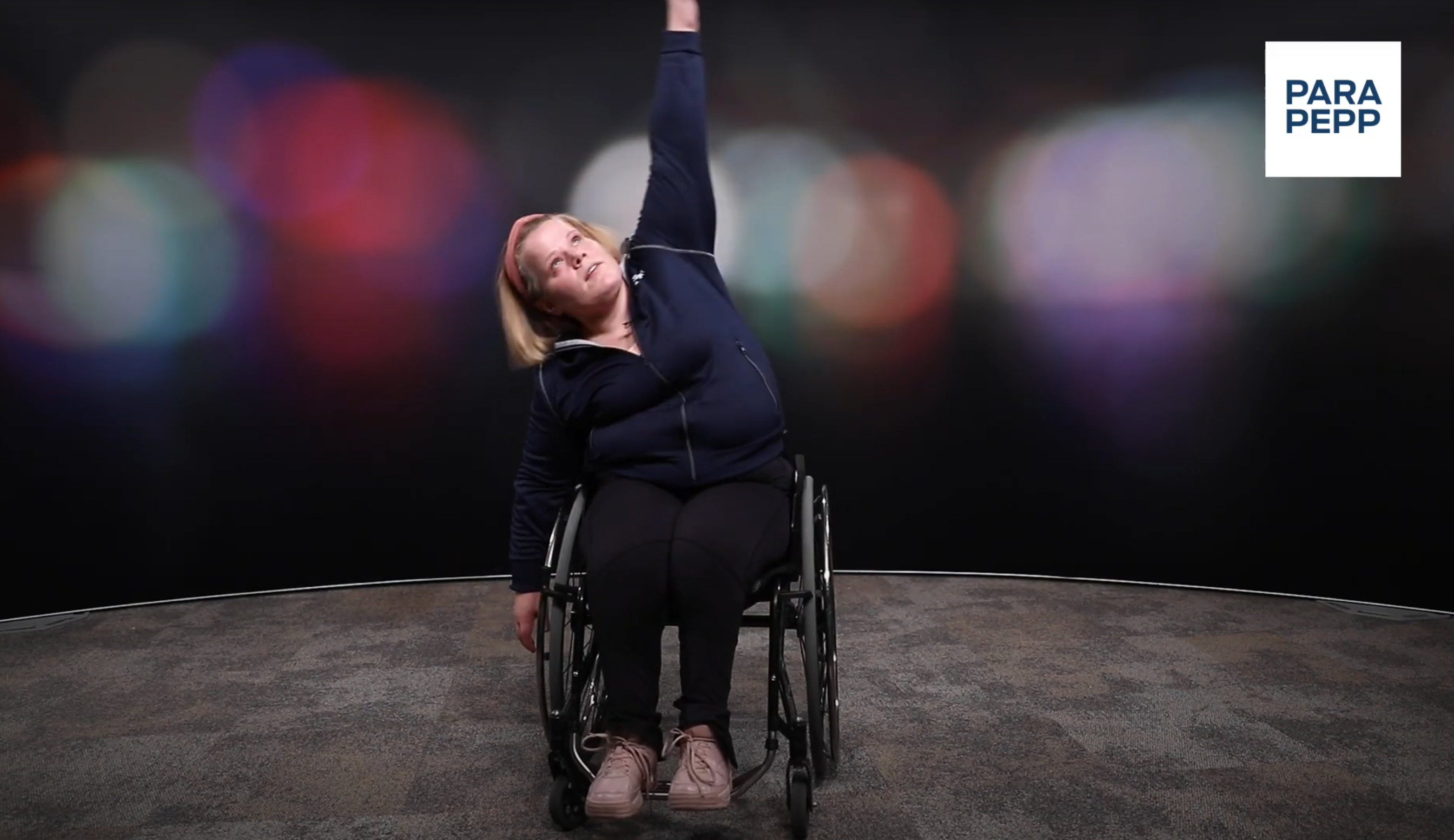 En kvinna sitter i rullstol. Hon har mörka träningskläder på sig och rosa skor. Hon sträcker vänsterhanden rakt upp, lutar sig till höger och låter vänsterarmen hänga.  Bakgrunden är mörk med suddiga cirklar i olika färger. 