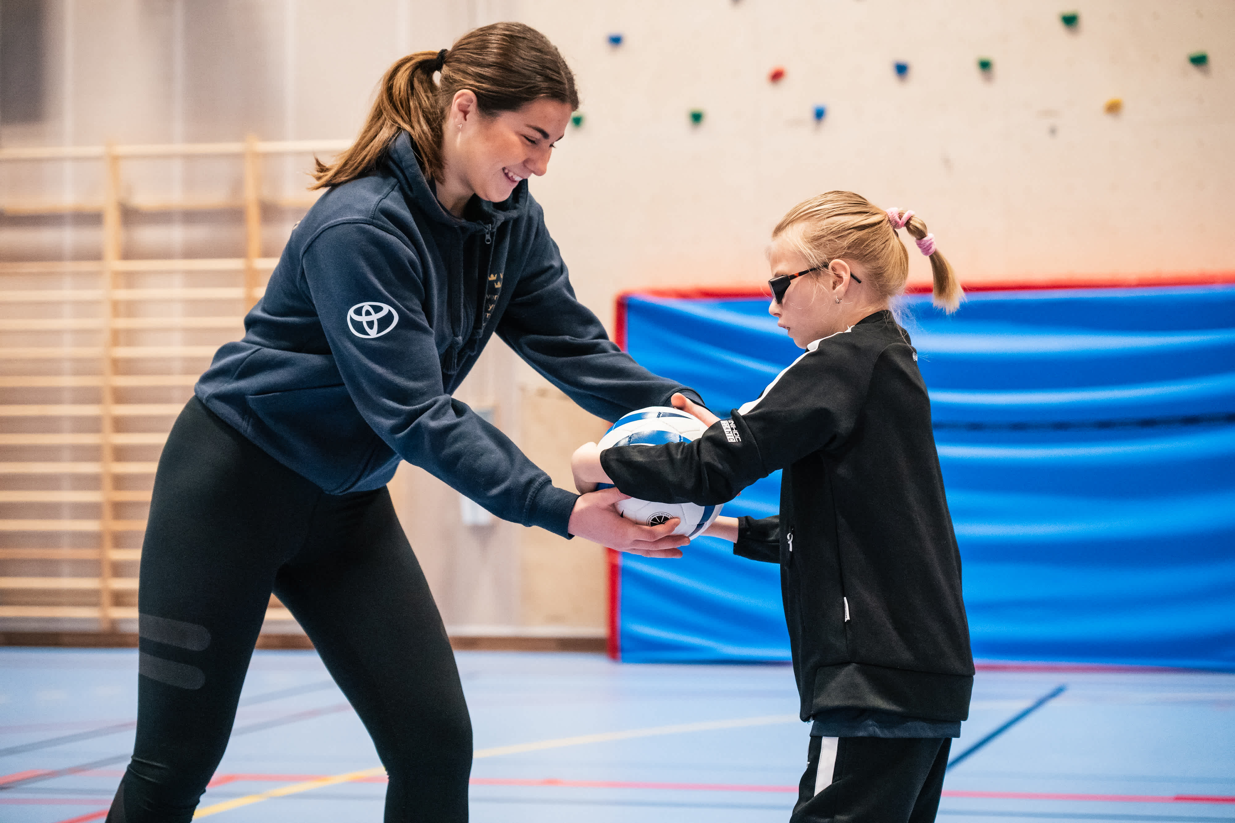 Inomhusmiljö, idrottshall. En kvinnlig ledare och en deltagare gör en övning där de båda håller i fotbollen med händerna.