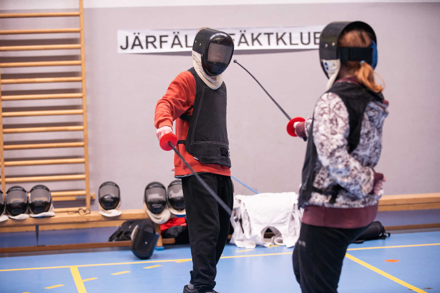 Två unga personer provar på fäktning i en idrottshall. Båda har varsin värja i höger hand och vänster hand bakom ryggen.