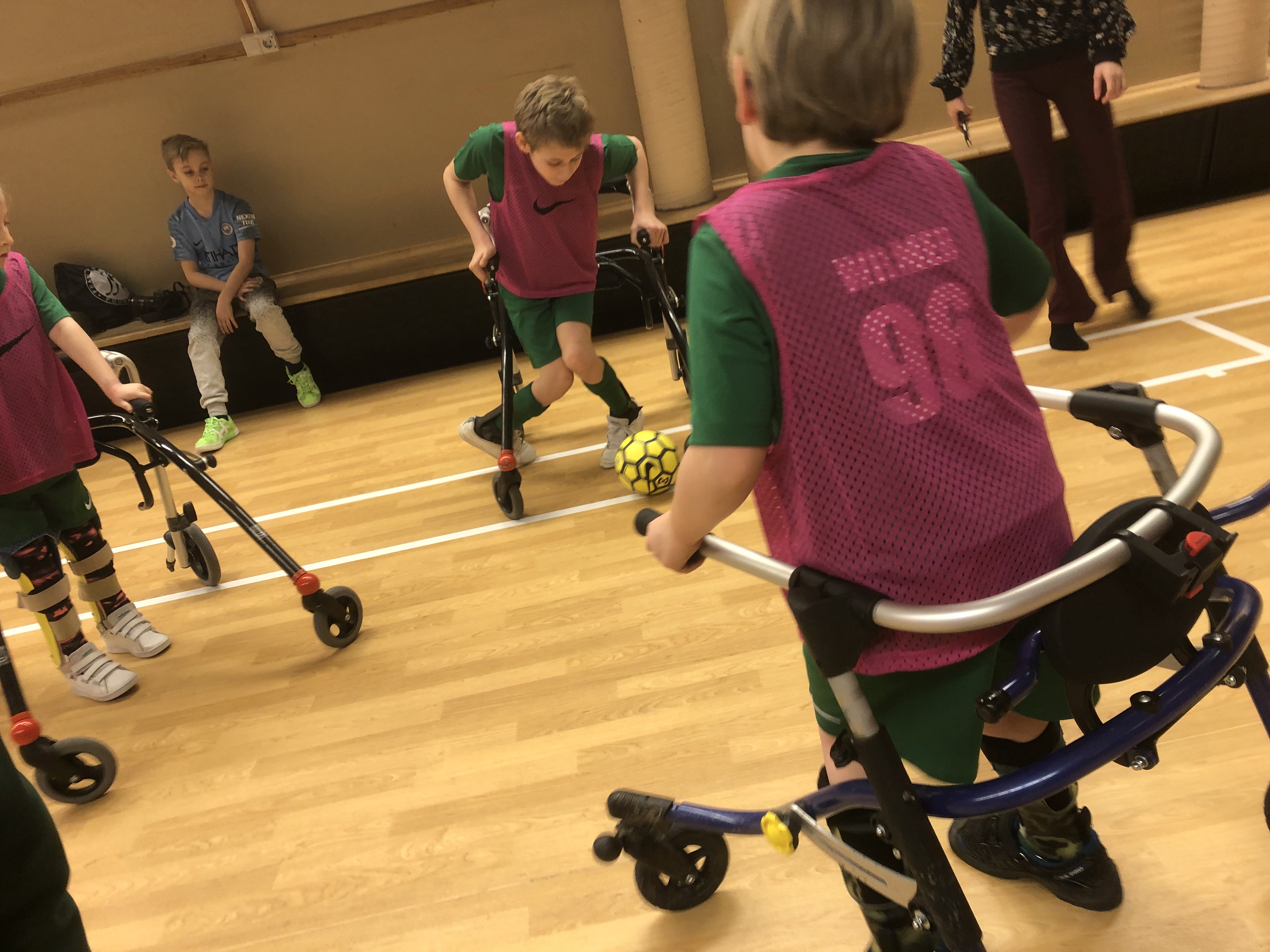 Tre barn spelar fotboll med bakvända rullatorer. En pojke har bollen vid sina fötter. De andra barnen kollar på.