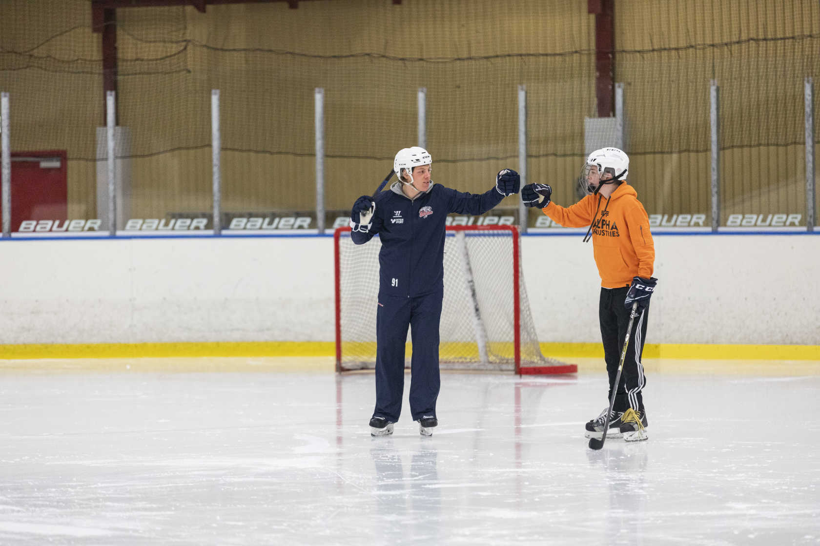 Två personer står på isen framför ett hockeymål. En av dem har en hockeyklubba. De slår ihop sina knogar i en hälsning. 