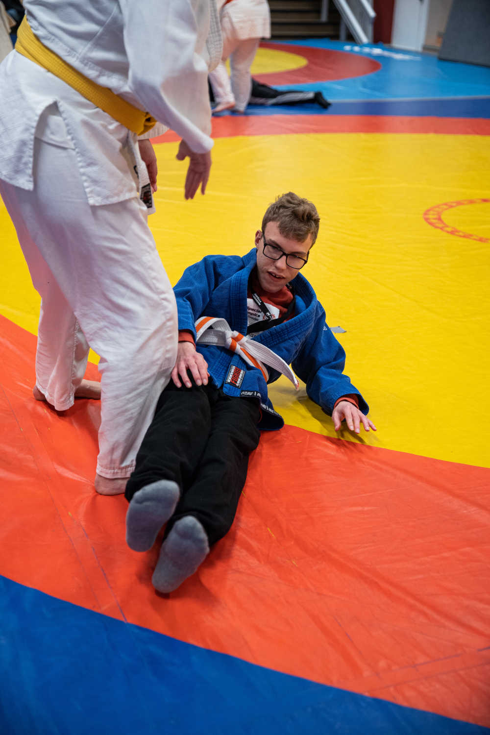 En kille tränar fall med judoinstruktören. Här håller han på att ta sig upp till sittande på mattan. Mattan är en cirkel med gult i mitten, sedan en röd cirkel och längst ut blått.