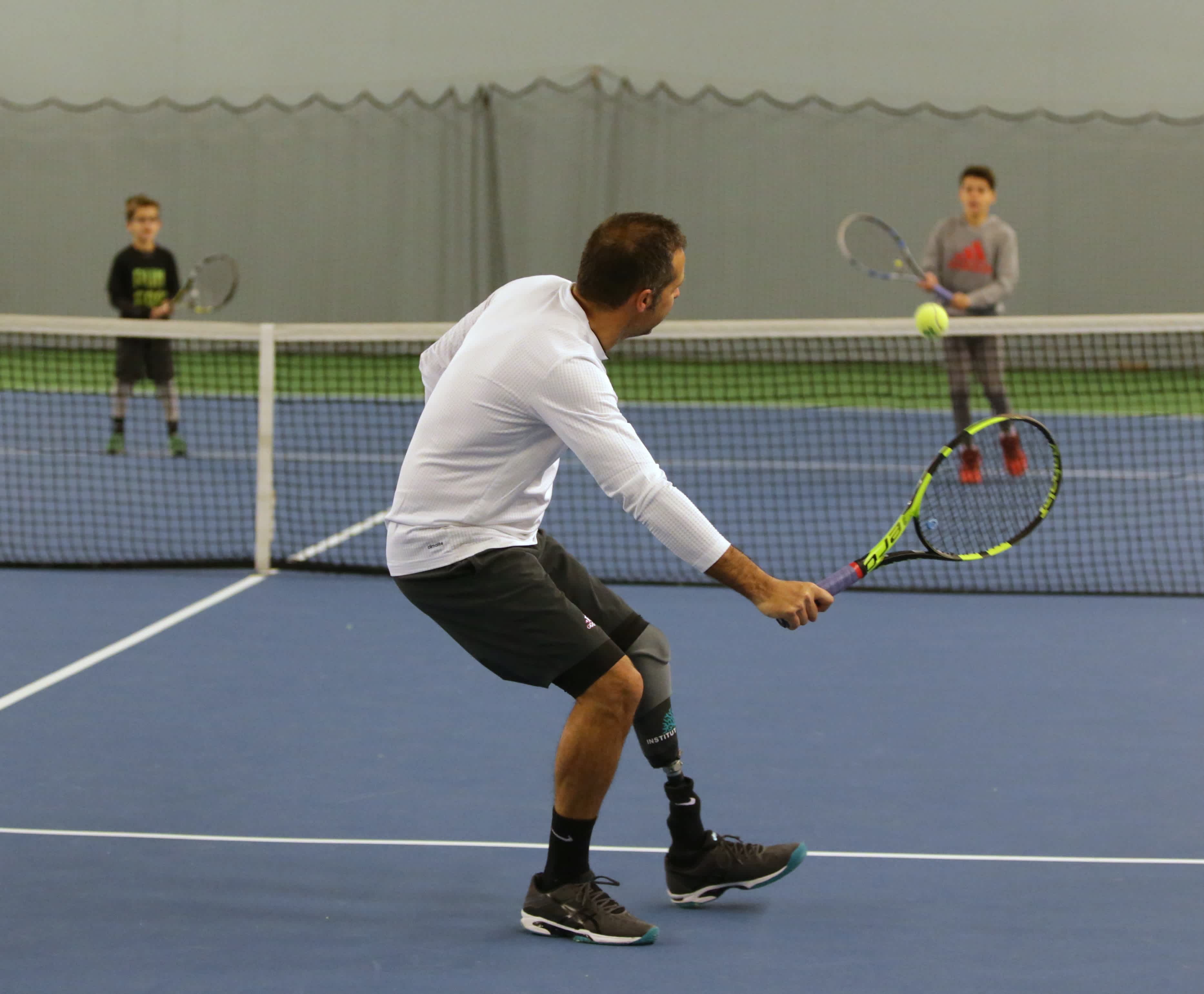 En man med benprotes spelar tennis mot två yngre pojkar.