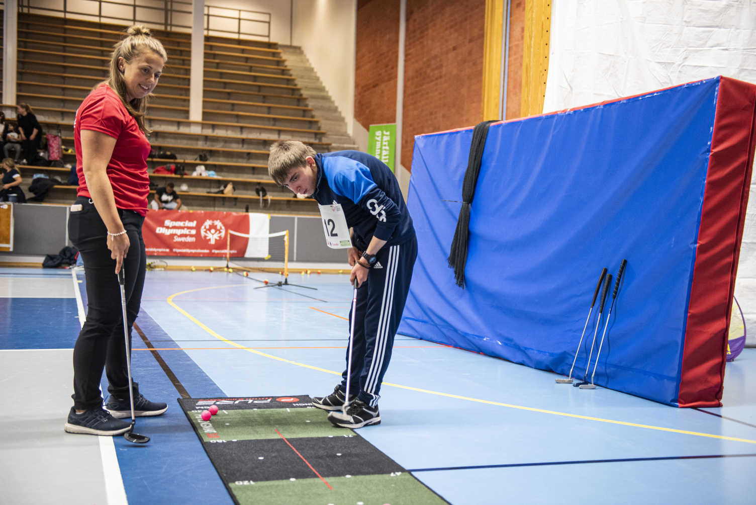 En kille spelar bangolf i en idrottshall och en kvinnlig instruktör tittar på. I bakgrunden står fler klubbor lutade mot väggen.