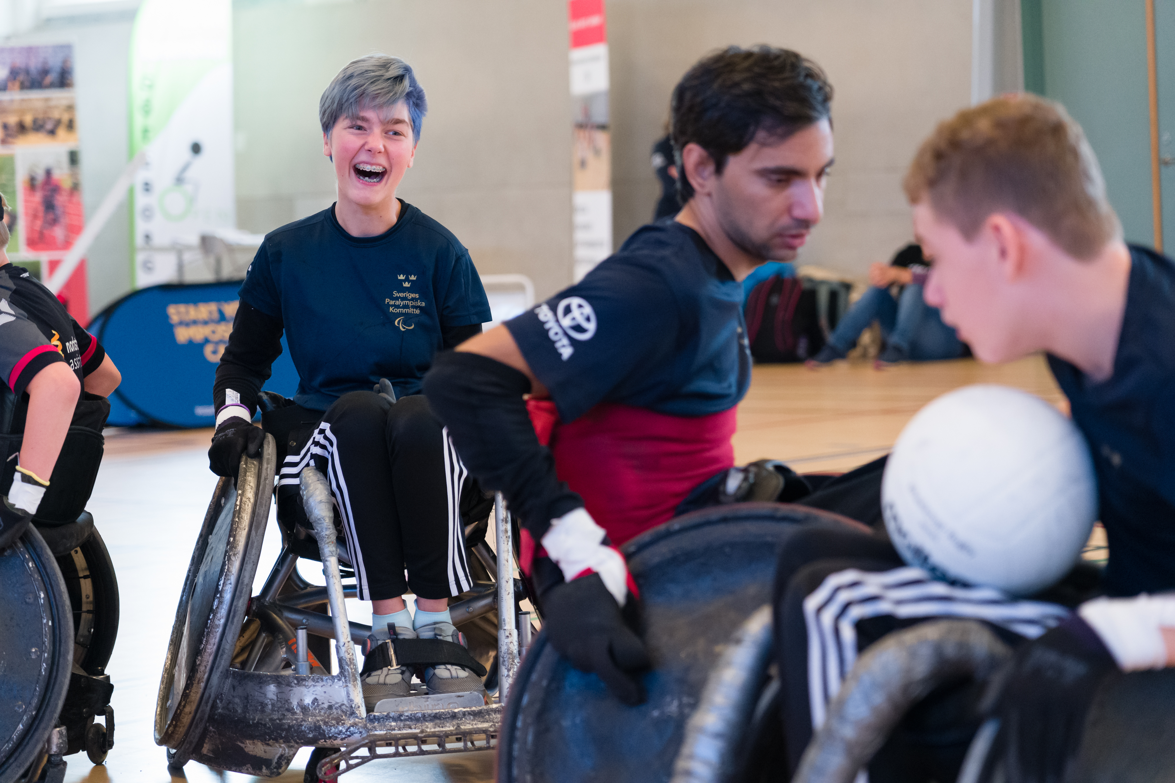 Tre rullstolsrugbyspelare. I förgrunden två killar som fajtas om bollen, i bakgrunden en tjej som ler stort. 