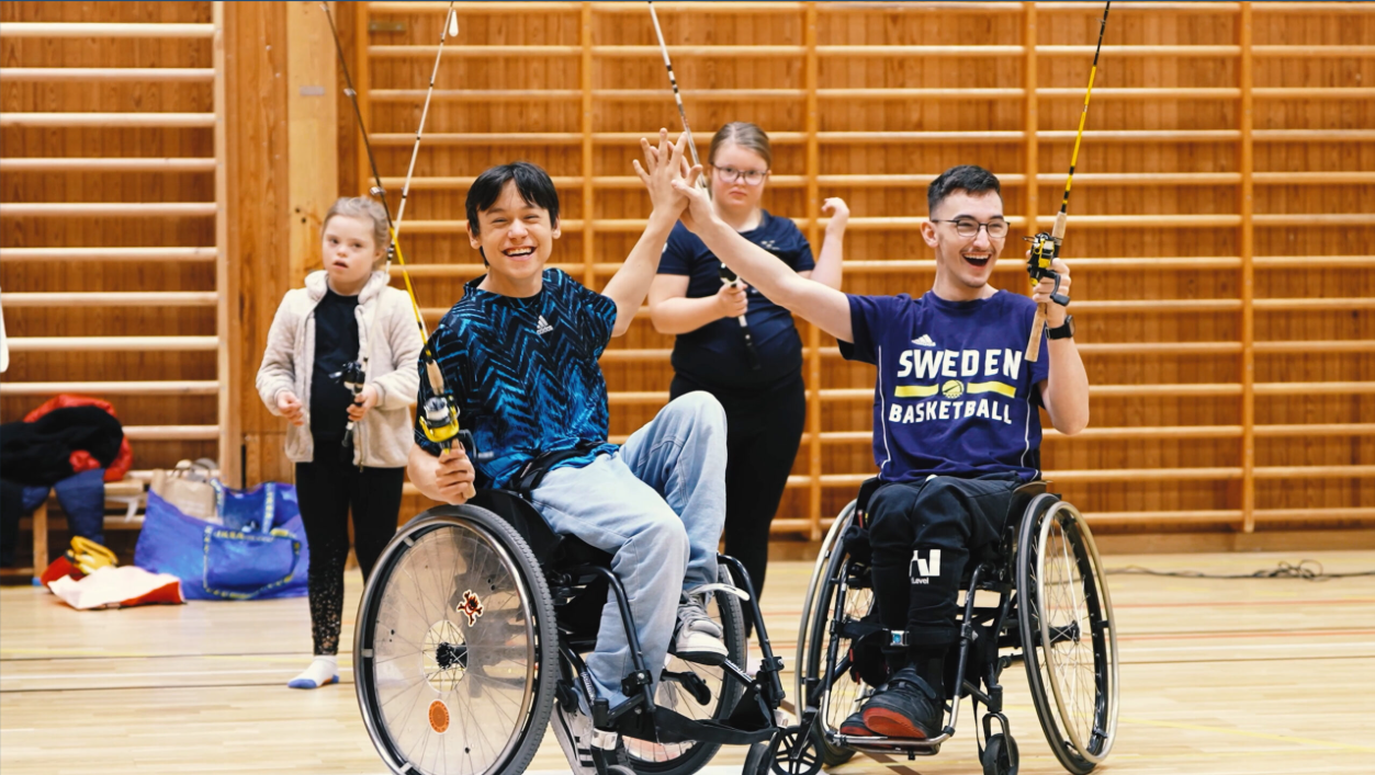 Inomhusmiljö, idrottshall. Två unga flickor och två killar i rullstol har alla varsitt spö. Det är glada och killarna gör high five.