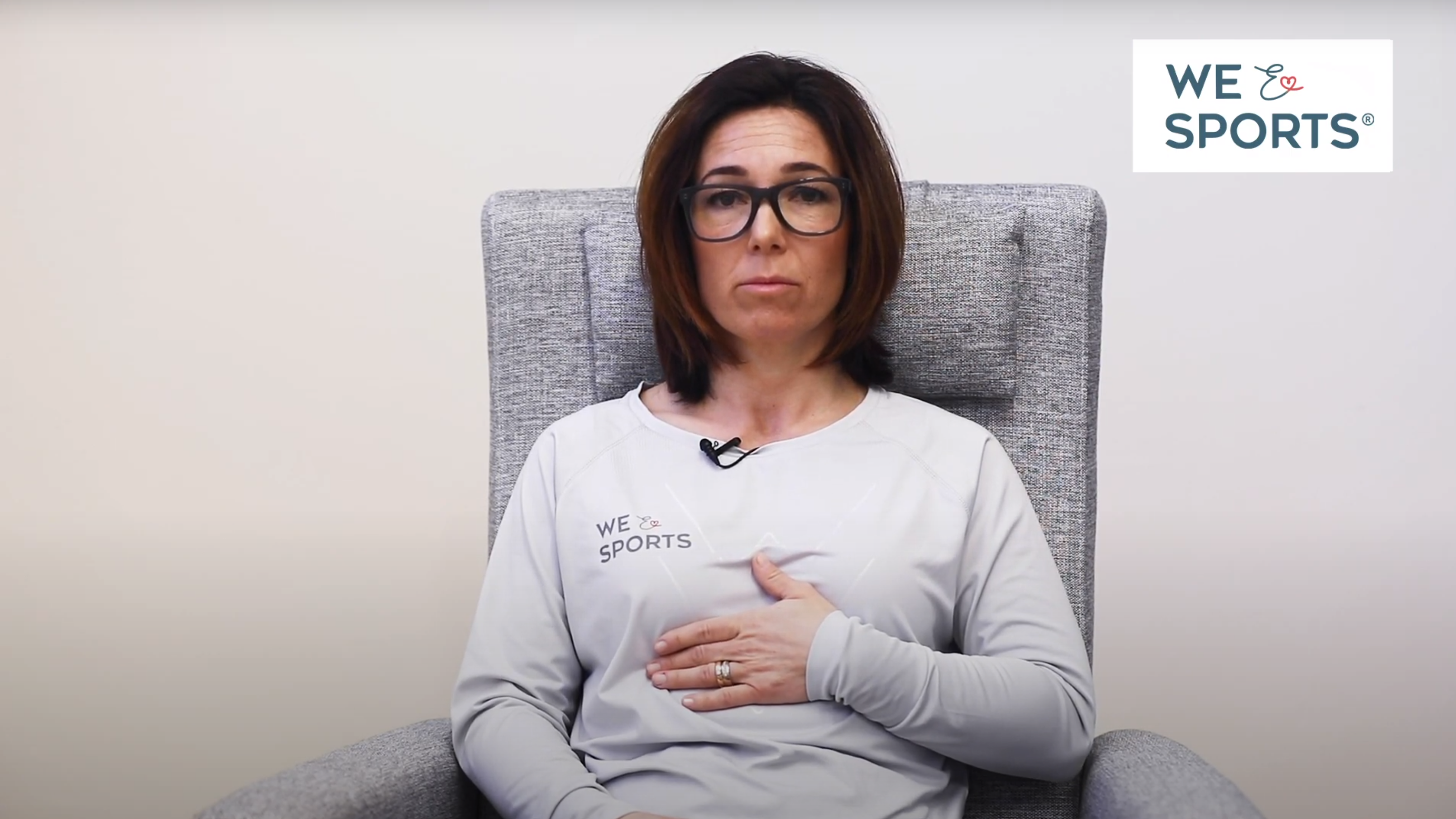 En kvinna med brunt, axellångt hår och svarta glasögon sitter i en ljusgrå fåtölj. Hon har ena handen mellan bröstkorgen och magen, för att demonstrera vart man ska andas ifrån. Hon har en vit, långärmad tröja på sig och bakgrunden är vit. 