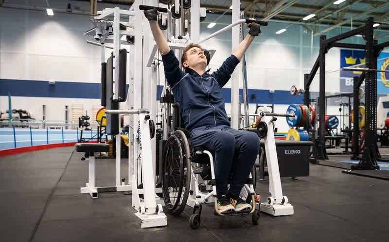 En man sitter i rullstol på ett gym. Han har blå tröja och blåa byxor. Han sitter vid en träningsmaskin och utför övningen axelpress. 