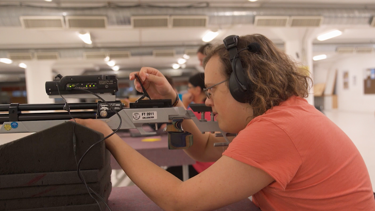 Inomhusmiljö, skyttehall. En ung man i profil. Han sitter vid ett bord med ett elektronskyttegevär i handen. Han har hörlurar.