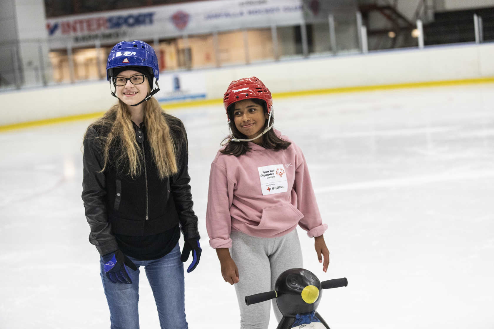 Två tjejer på isen. De har hjälm på huvudet samt stöd för att hålla balans.