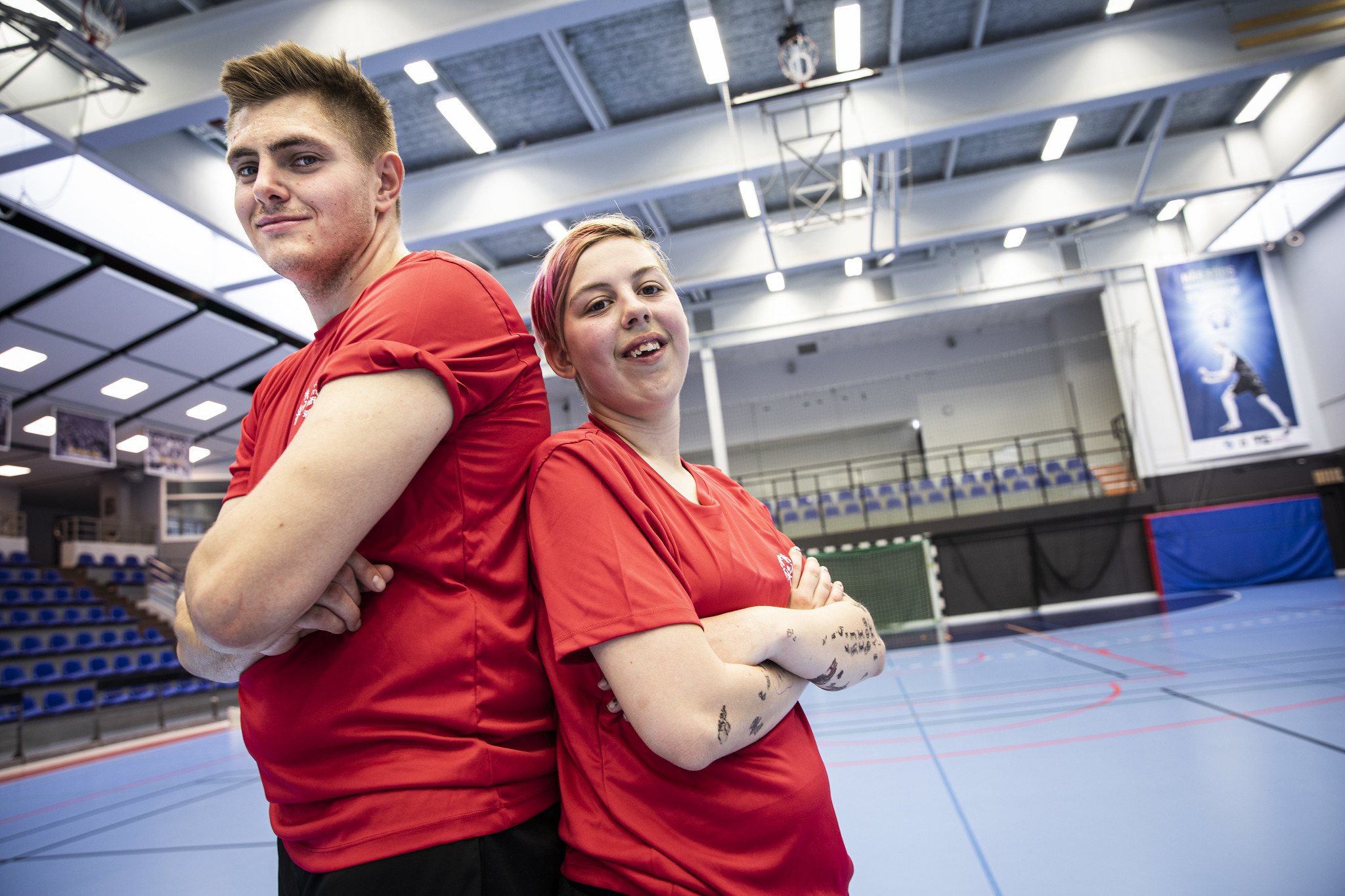 Ricky (man) och Alva (kvinna) står rygg mot rygg med armarna i kors. De har röda t-shirts och i bakgrunden är en stor idrottshall med blått golv och streck i olika färger. Det är läktare på ena kortsidan och ena långsidan. På andra sidan hallen syns ett fotbollsmål och en tjock idrottsmatta.