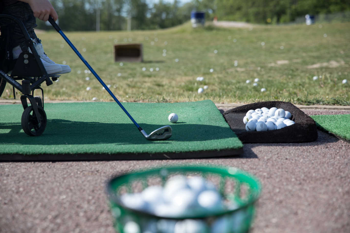 Utslagsplats vid golfbana, i förgrunden står en hink full med golfbollar, person i rullstol förbereder sig för att slå. 