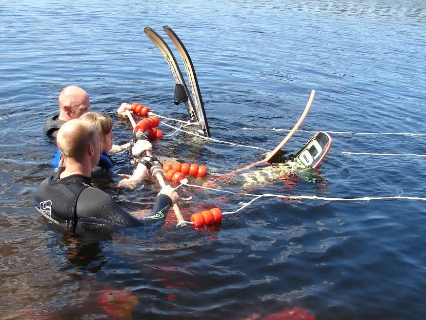 Tre personer med vattenskidor på fötterna flyter i vattnet och håller i en tredelad bom som sitter fast i en lina.