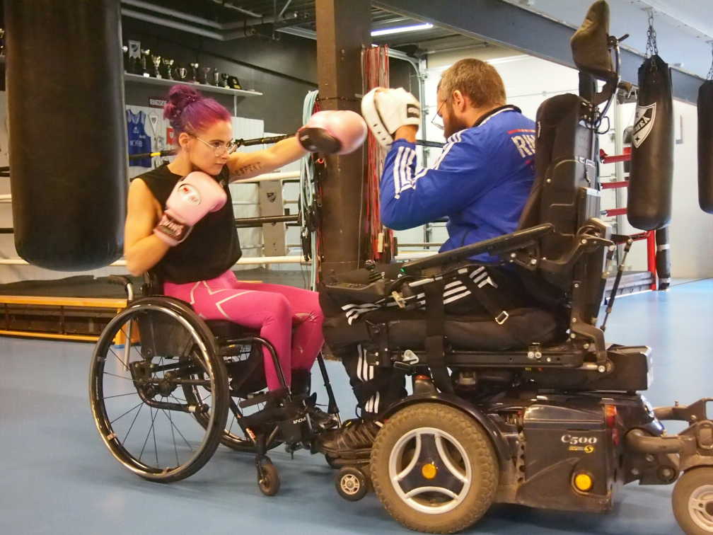 Inomhusmiljö, boxningslokal. Två personer i rullstol tränar boxning med varandra.
