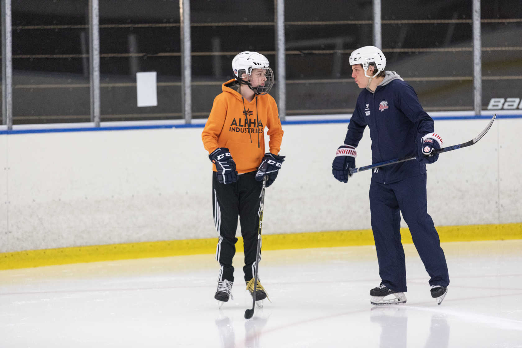 Två personer åker skridskor på isen och pratar med varandra. De har varsin ishockeyklubba.