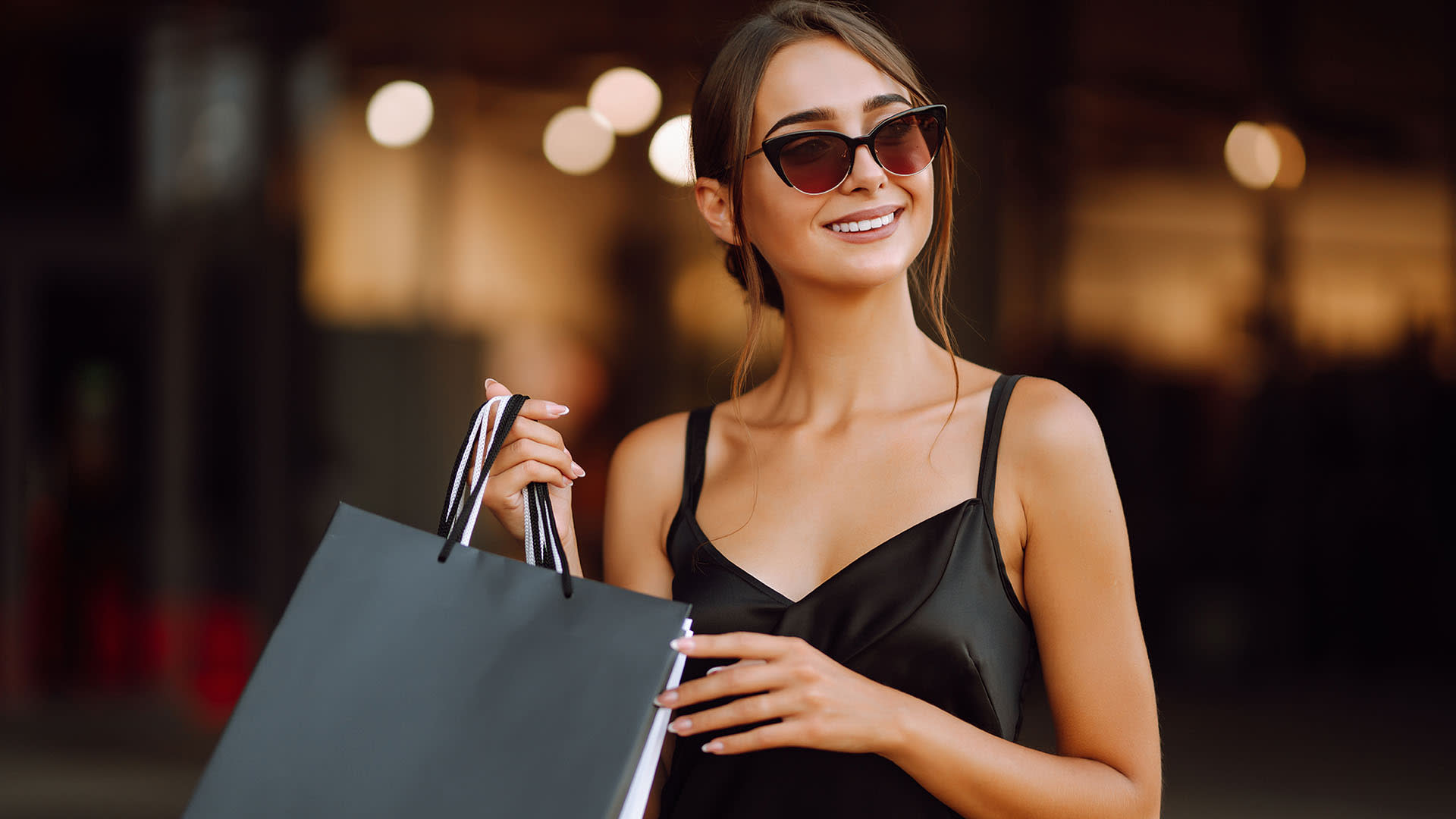 Sugar Baby - Uma mulher jovem e atraente. Uma sugar baby, mulher morena, sorrindo e usando óculos escuros, segura sacolas com presentes dados pelo seu sugar daddy.