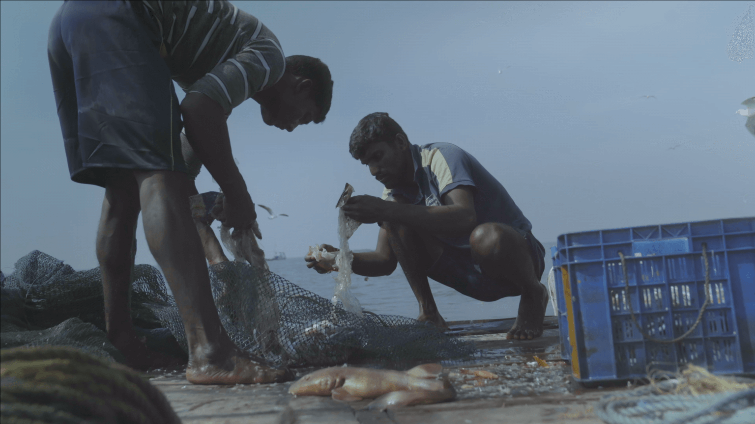 Film still from Against the Tide. Two men go through the content of their fishing net, there are a fish and plastic trash.