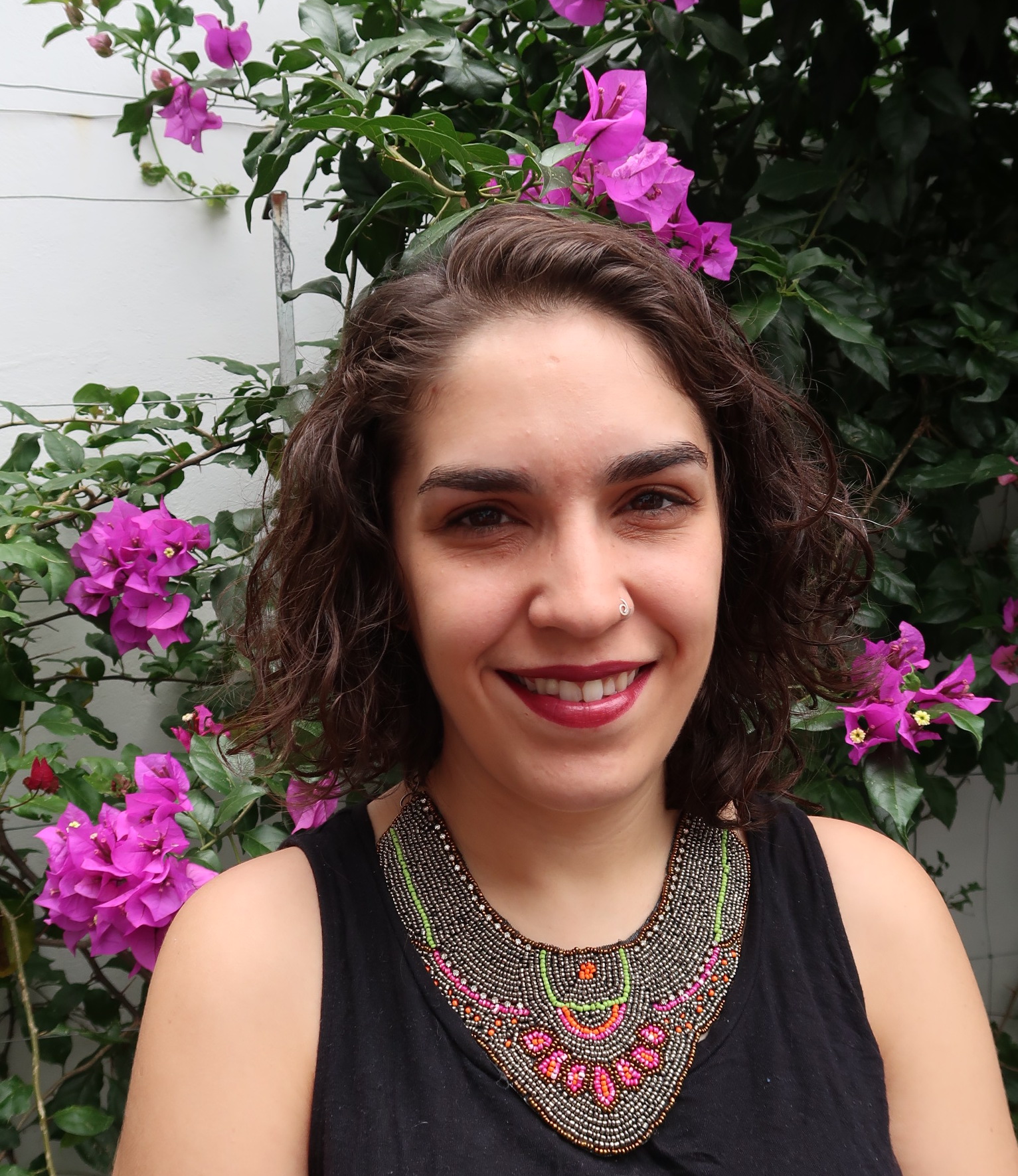 Headshot of Director Indira Cato, a woman with curly medium hair, smiling at the camera, on a garden with purple flowers.