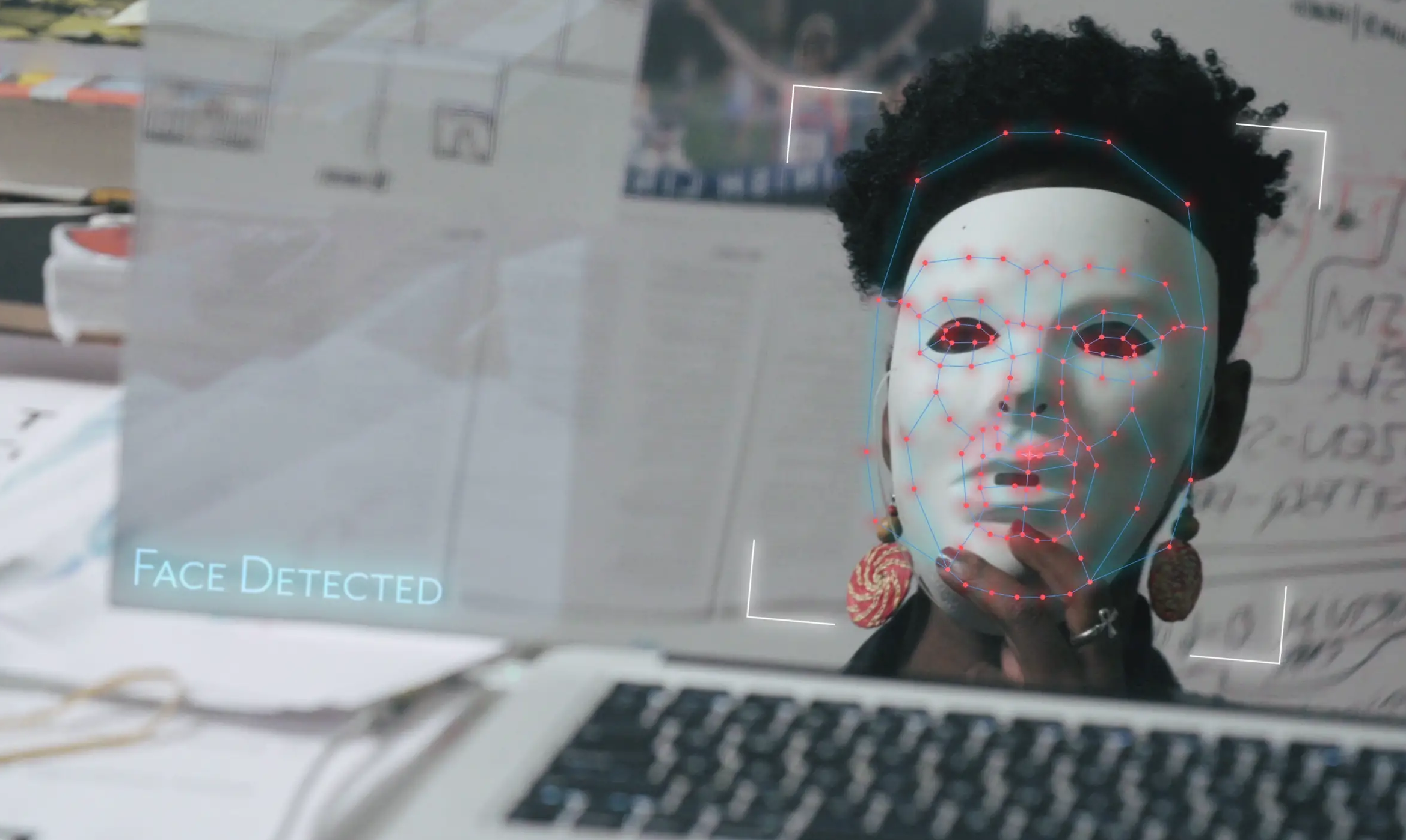 Still from Coded Bias. A woman holds a white mask up to her face and facial recognition software writes 'face detected.'