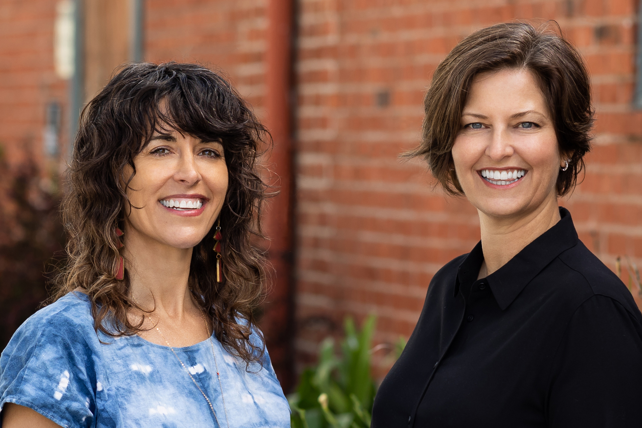 Two women are pictured together standing outside in front of a brick wall.
Left: Rivkah, a white woman with shoulder length curly hair stands in a blue and white blouse smiling. 
Right: Jen, a white woman with short brown hair, stands in a black top smiling. 