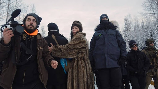 A group of people stand in a line shielding each other from impending violence. A person holds a camera and police in military garb circle. 