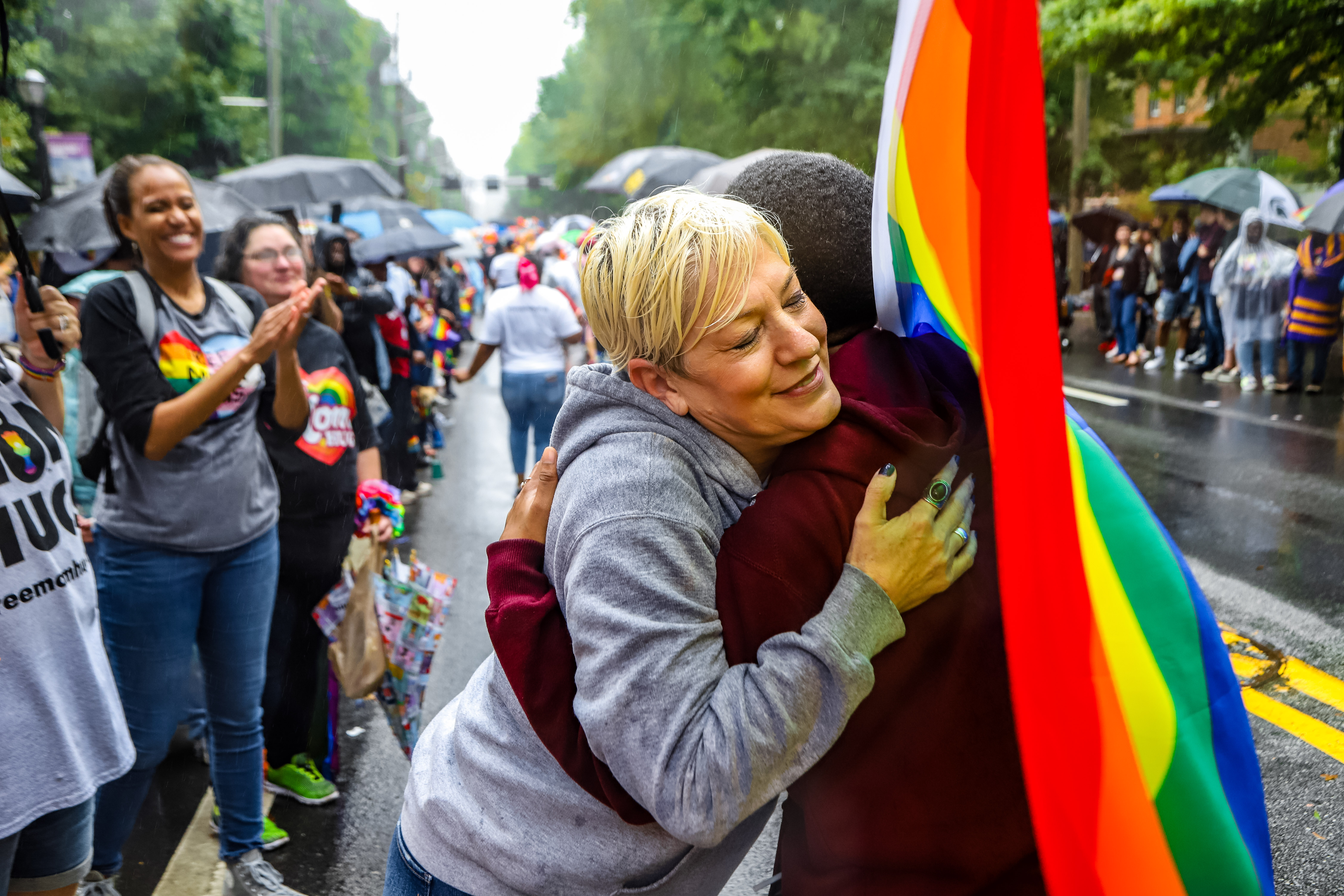 Two people hug and smile at a pride rally in the rain. They are covered by a gay pride flag and are being cheered on by people on the sidelines.