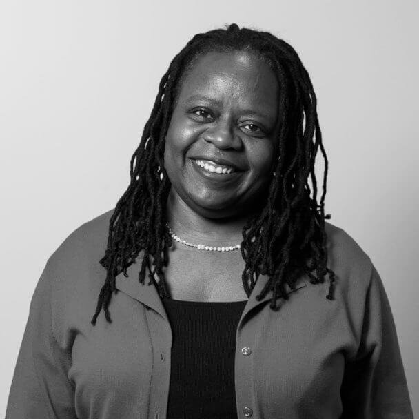 Headshot of a woman tilting her head to her left and widely smiling. She wears dreadlocks, an open shirt with a shirt underneath and a necklace.