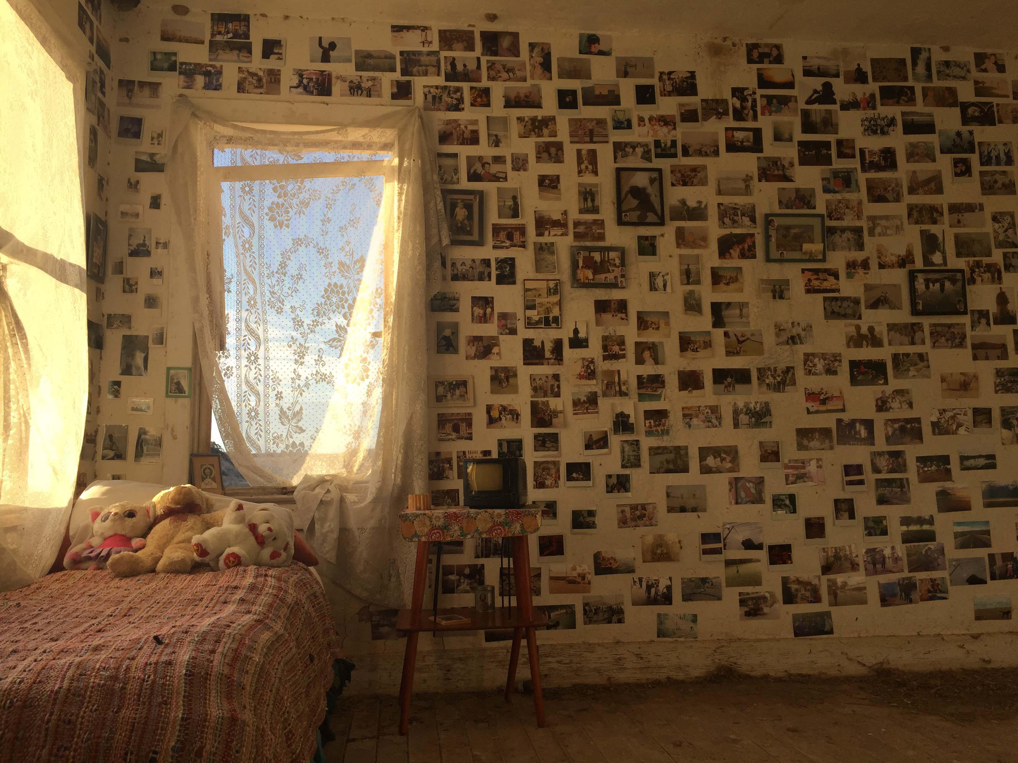 Still from The Guardian of Memory. The interior of a bedroom. One wall has a window and the rest of the wall is covered in photos.