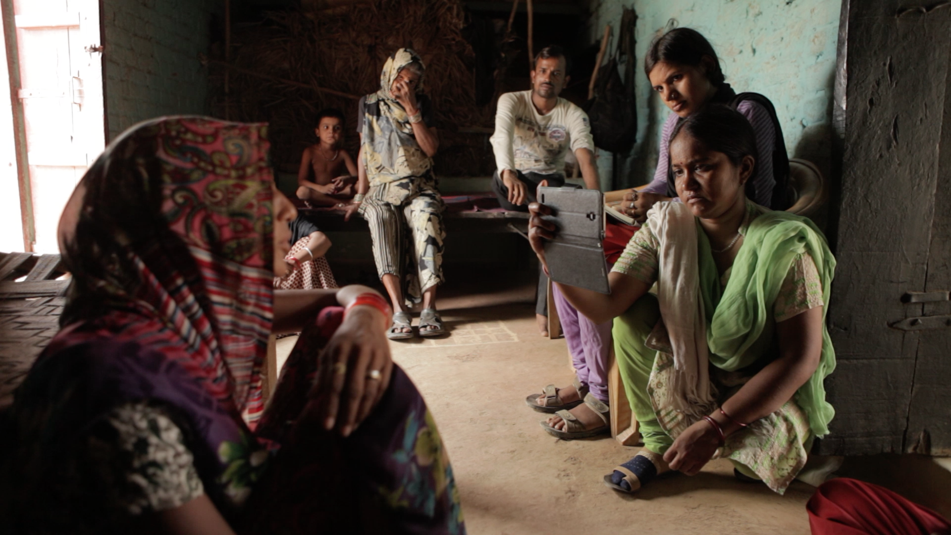 Still from Writing With Fire. Two women sit on the ground, one holding up a phone in a gray case. She records a woman wearing a colorful, patterned hijab and pants. In the back, a woman in a hijab, a man, and a young boy are sitting on a bed.