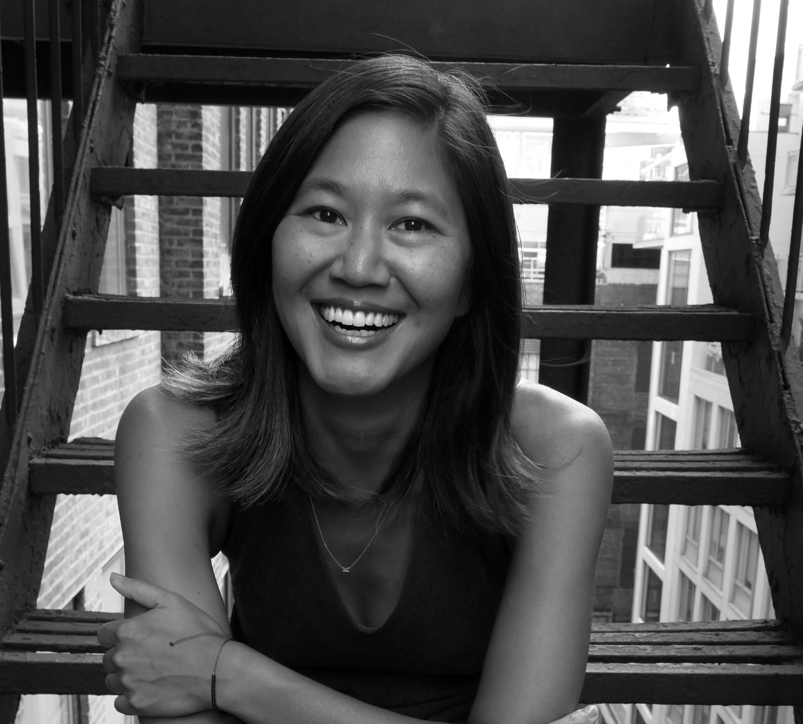 Stephanie Wang-Breal sitting on fire escape stairs, smiling widely. She has mid-shoulder length, dark hair, and wears a sleeveless shirt. Black and white portrait.