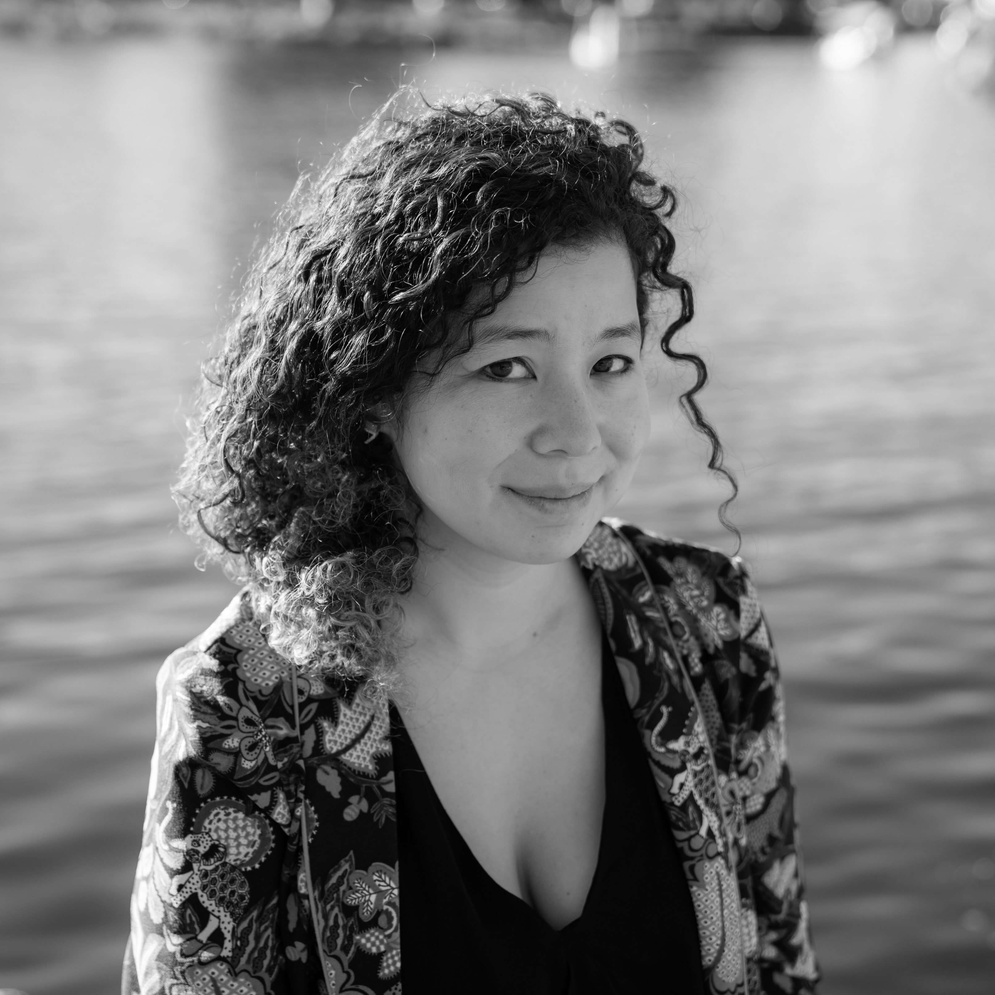 A headshot of filmmaker Jessica Kingdon. She sits in front of a lake, wearing a black v-neck shirt and a floral printed blazer. Her curly hair is pulled over her right shoulder. She looks at the photographer/viewer smiling. 