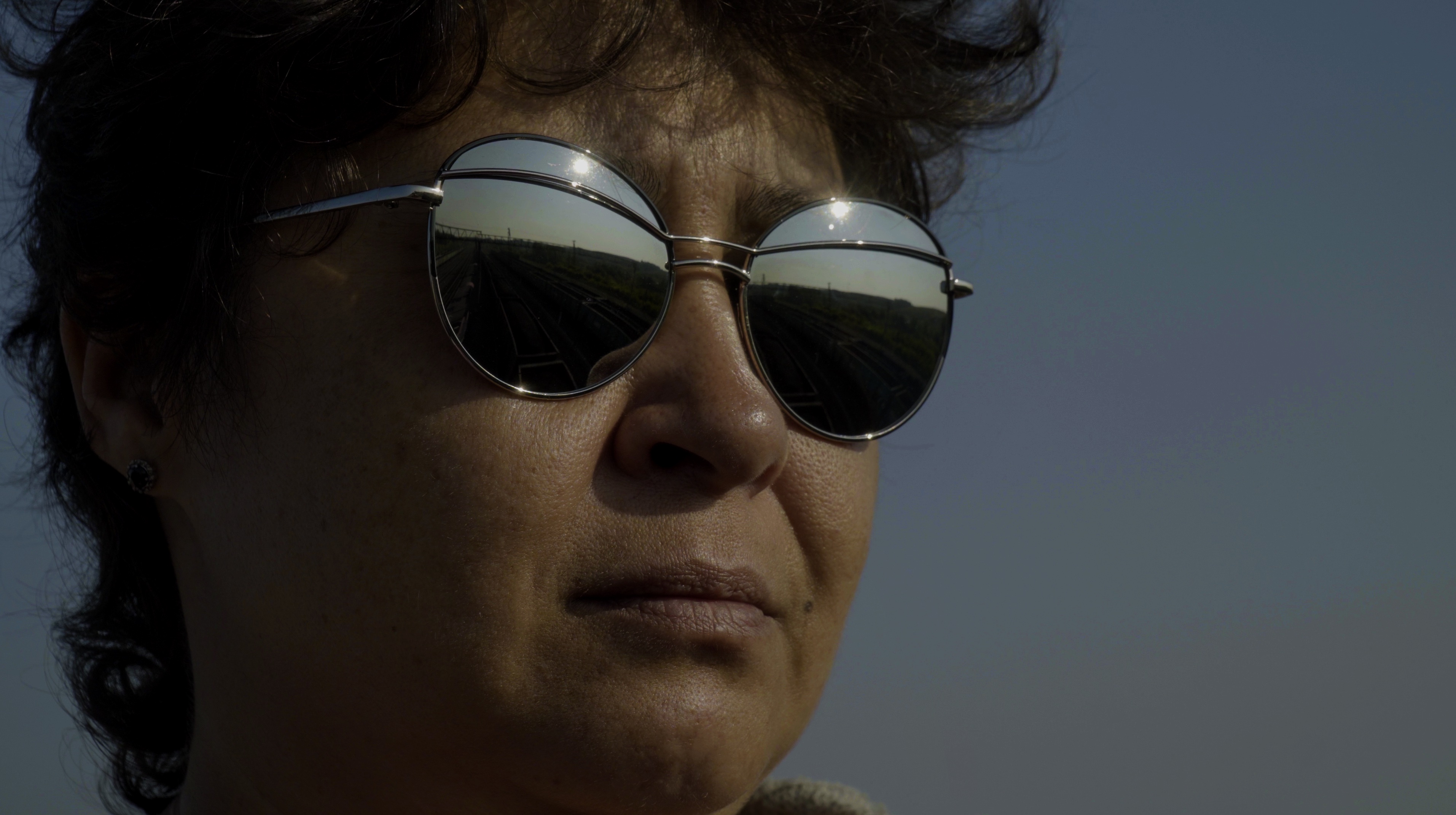 Still from Black Snow. A head-only view of a woman with short, brown hair wearing mirrored sunglasses with coal trains reflected in them.