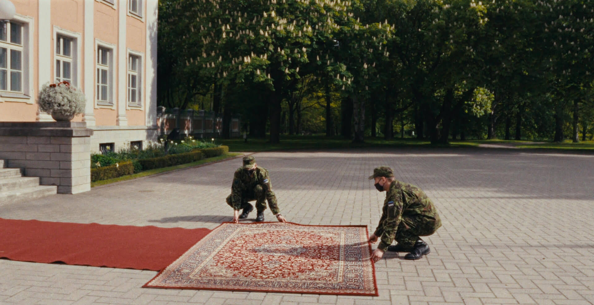 Still from The Wife Of. Men in military uniforms straighten a rug in a driveway.