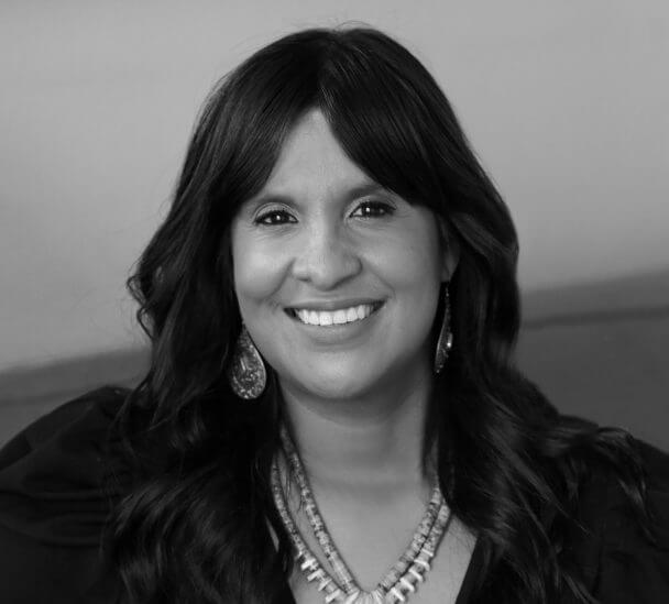 A woman with dark skin, long, wavy black hair and black eyes sits on a bright red/orange sofa, her hands resting on the sofa's arm rest. She's smiling while looking into straight into the camera. She is wearing a long sleeve black dress with a V-neck,has on a turquoise necklace and matching earrings.