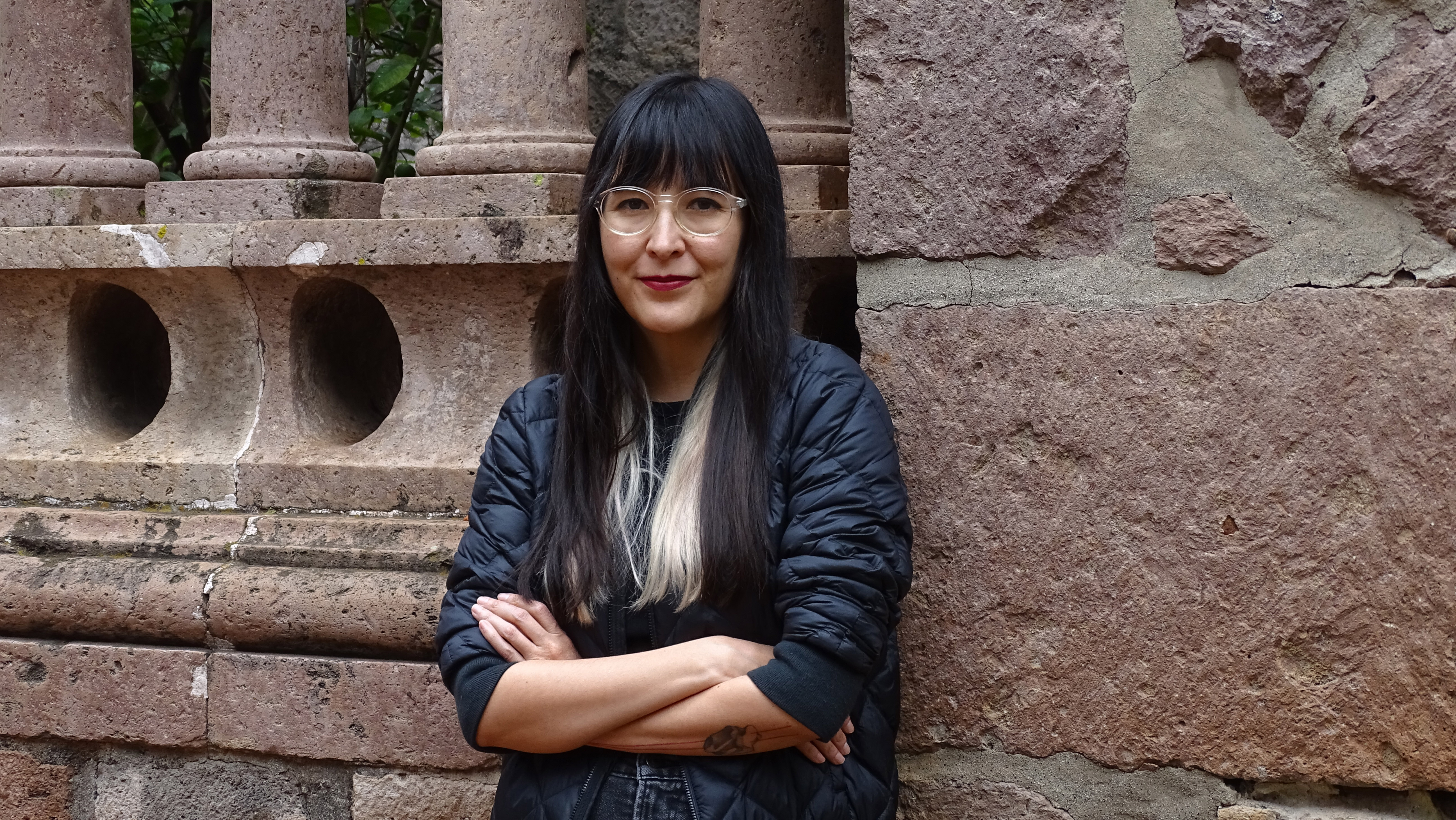 Headshot of Sumie García smiling at the camera wearing black clothing, arms crossed and leaning on a wall made of reddish stone.