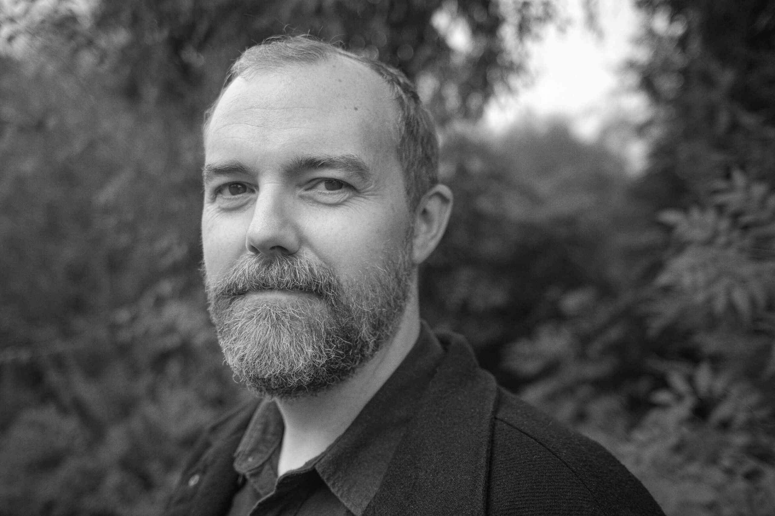 A middle aged man with a full beard and short hair looks over his left shoulder at the camera. He wears a dark collared shirt and dark suit jacket. Headshot of director - producer Joe Peeler of Bad Press. 