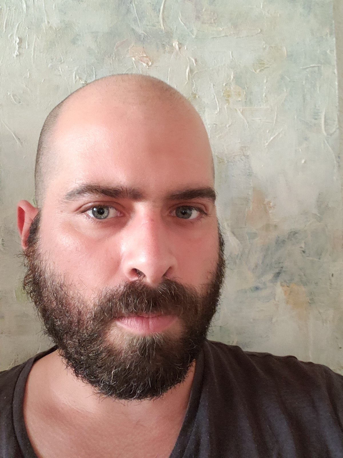 Close-up headshot of Carlos Paz, looking directly at the camera, wearing a black t-shirt in front of a splotchy off-white background.