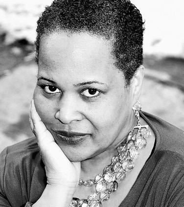 Ann Bennett looking directly at the camera. She is wearing a necklace and earrings and is placing her hand against her face. Black and white portrait. 