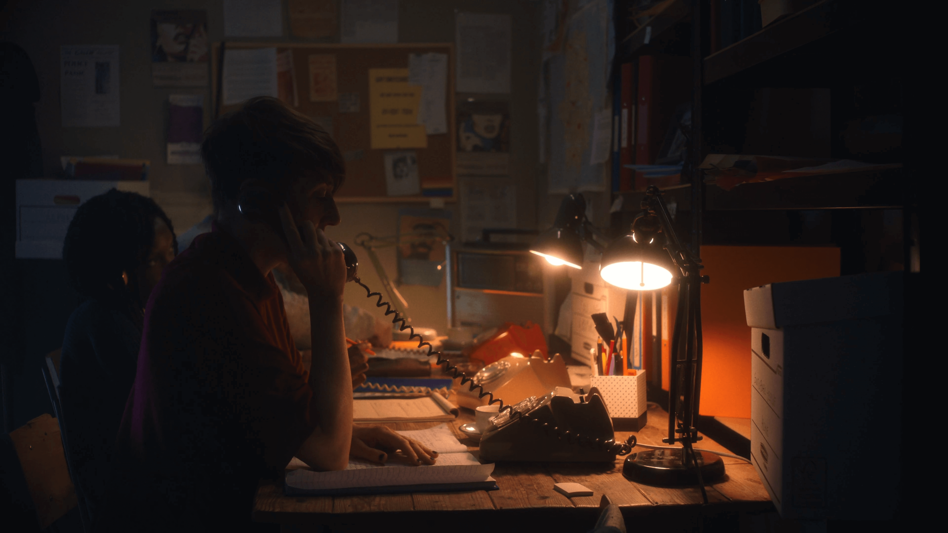 Still from The Callers. In a dimly-lit, messy office, two people sit at a desk speaking on rotary phones.